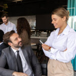 Smiling businesswoman in a white shirt talking with a seated colleague in a suit at a restaurant table