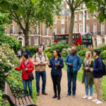 Tour guide leading a small group with audio headsets through a London garden square, red double-decker bus behind