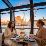Two women enjoying afternoon tea with scones and cakes on a Thames river cruise, the London Eye behind them