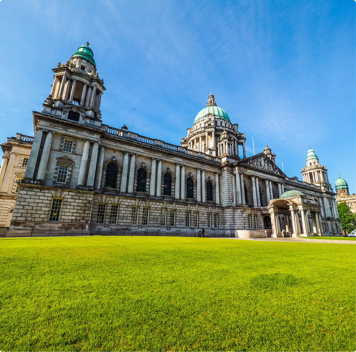 Belfast City Hall with its green copper domes and stone facade behind a manicured lawn on a sunny day