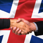 Two people in dark suits shaking hands in front of a Union Jack flag