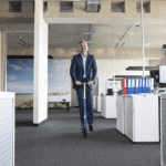 Smiling businessman in a navy suit riding a kick scooter through a modern open-plan office