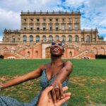Smiling woman in sunglasses reaching back to hold a companion's hand on the lawn in front of Cliveden House in Buckinghamshire