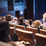 Audience in an auditorium photographing a speaker at a podium during a conference presentation