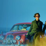 Smiling couple in vintage-style clothing walking hand in hand past a classic red car in the countryside