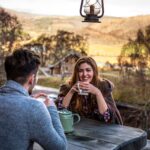 Smiling woman wrapped in a tartan blanket sharing tea with a companion on a log cabin deck overlooking an autumn valley