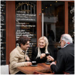 Three older friends chatting over espresso at an outdoor table in front of a French bistro with a hand-painted menu