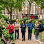 Tour guide with a headset speaking to a small group wearing audio receivers in a London garden square, red double-decker bus behind