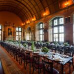 Long banquet tables set for a formal dinner in a historic great hall with a vaulted wooden ceiling, stained-glass windows and candelabras