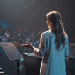 Woman speaking at a podium with teleprompters on a conference stage, addressing a large audience in a dark auditorium