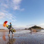 Kitesurfer and windsurfer carrying their boards across the beach at low tide, with St Michael's Mount in Cornwall behind