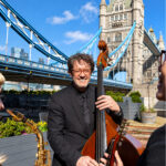 Smiling double bass player performing with a jazz band on a riverside terrace beside Tower Bridge in London