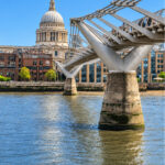 Steel supports of the Millennium Bridge rising from the River Thames, with the dome of St Paul's Cathedral behind on a sunny day