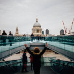 Visitor raising their arms in celebration at the foot of the Millennium Bridge, with St Paul's Cathedral ahead on an overcast day
