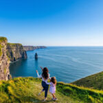 Mother and daughter on a grassy clifftop looking out over the Cliffs of Moher and the Atlantic Ocean on a sunny day