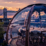 Glass dining igloo with a table set for dinner on a Thames riverside terrace, Tower Bridge behind at sunset