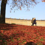 Couple walking across a lawn covered in autumn leaves in front of the Royal Crescent in Bath
