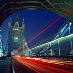 Tower Bridge at night with red and white light trails from passing traffic, framed by the arch of one of its towers