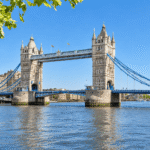 Tower Bridge spanning the River Thames on a clear summer day, framed by green leaves in the foreground