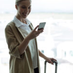 Smiling woman checking her phone while holding a suitcase handle by an airport terminal window